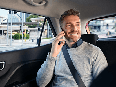 Smiling man in a grey sweater talking on a smartphone in a car's backseat.
