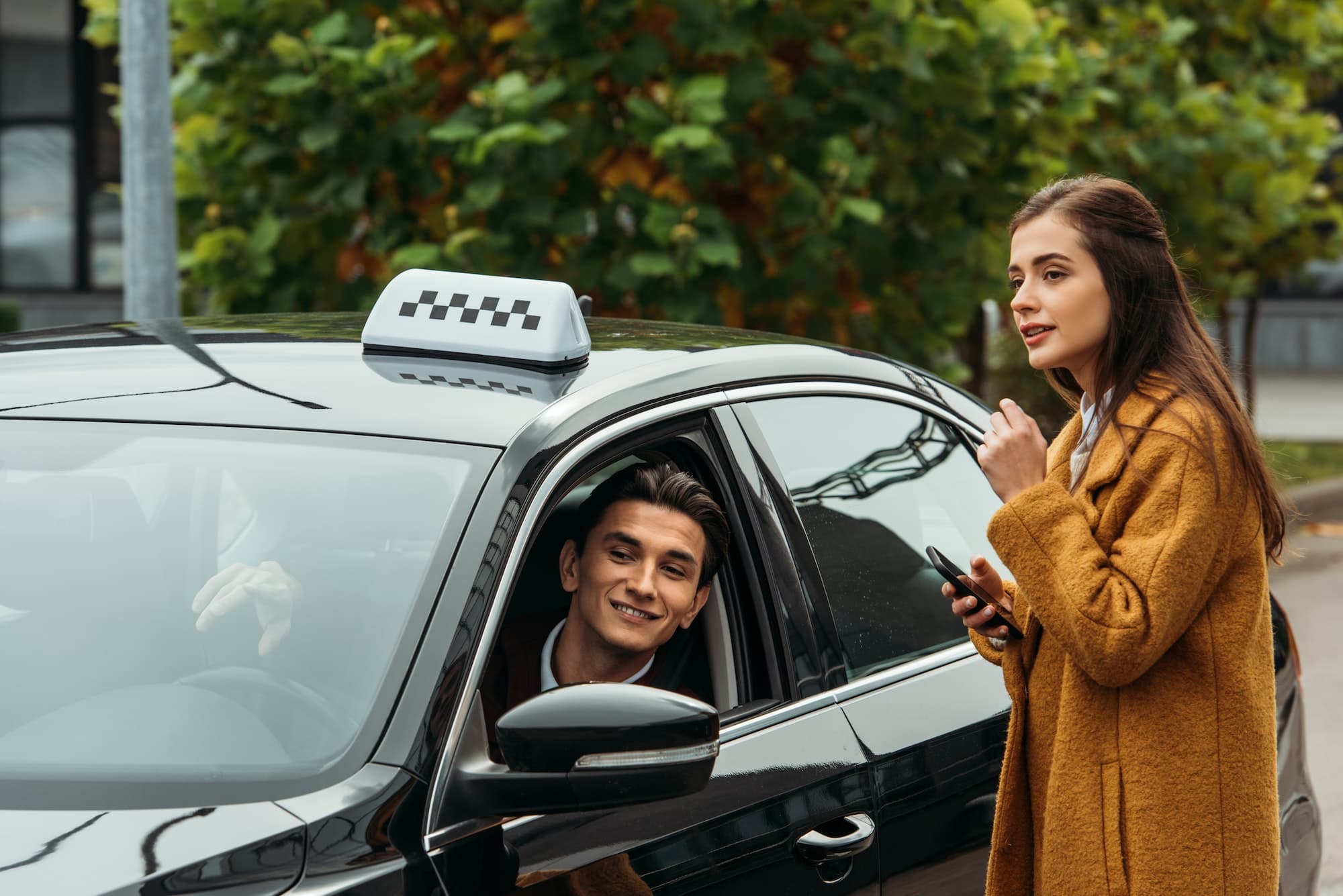 Woman in yellow coat talks to a smiling taxi driver through the car window.
